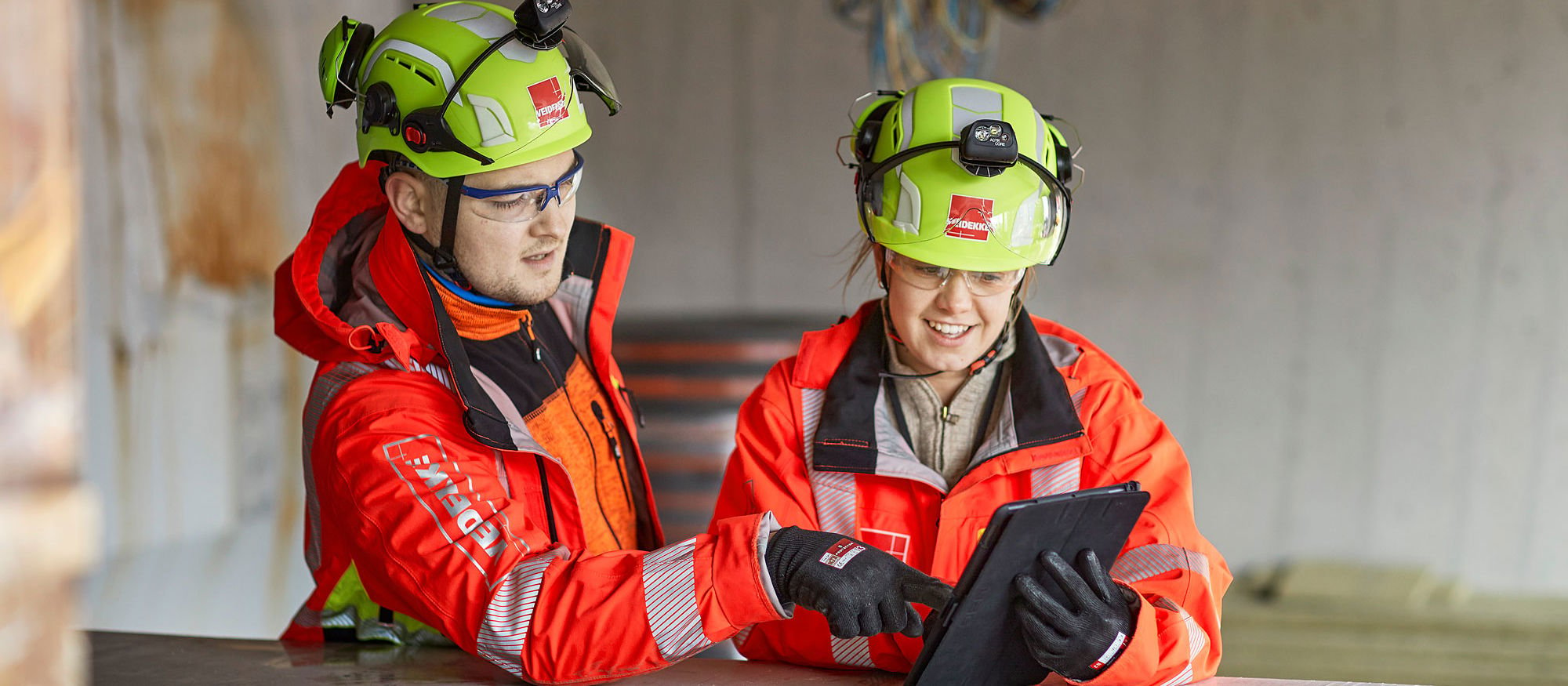 Two Veidekke employees looking at an ipad. They are wearing helmets and high-vis clothing. Photo.