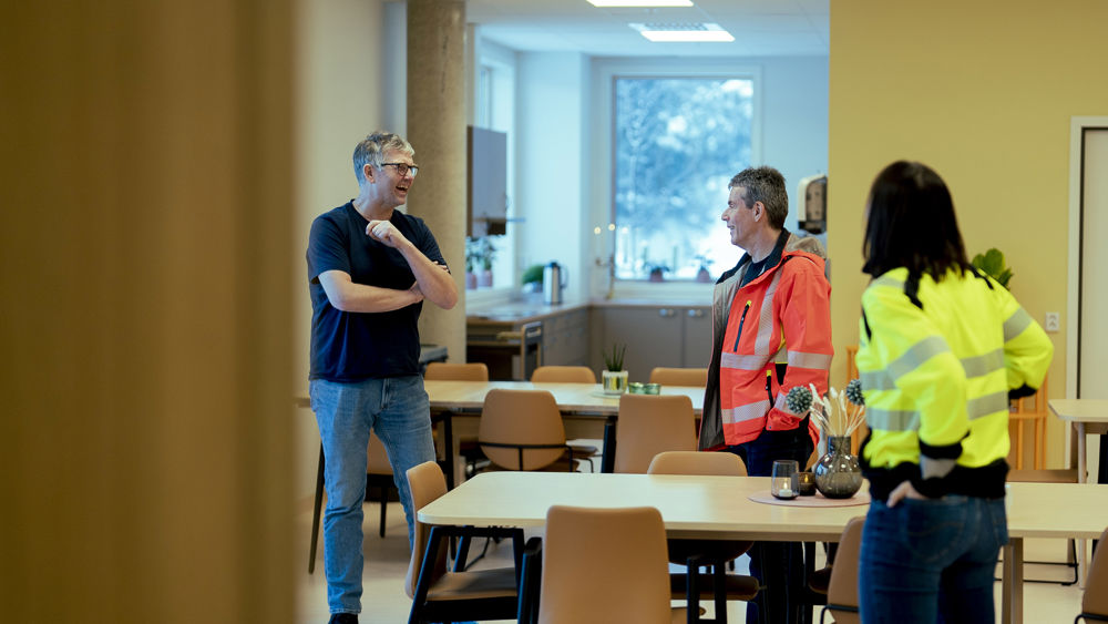 Tre personer som står inne i et rom og prater sammen. Foto.